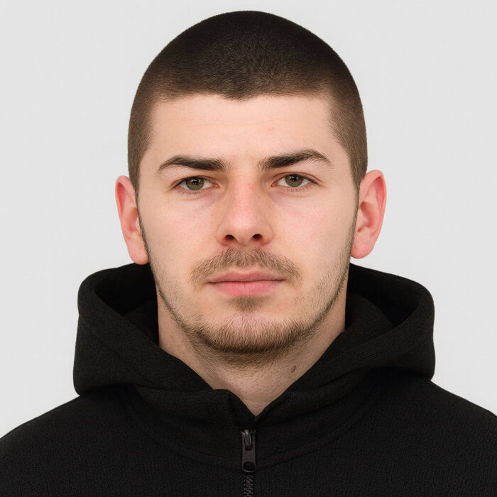 Млад мъж в черна качулка с сериозен поглед и неутрална изражение.