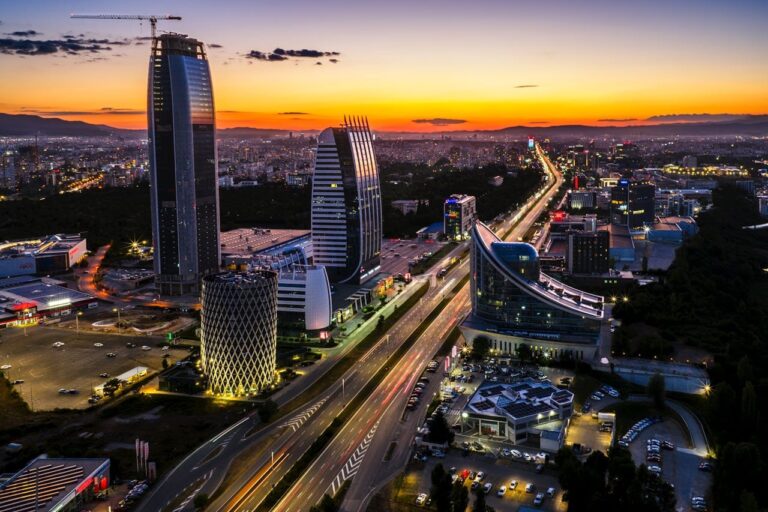 Модерен градски пейзаж при залез с високи сгради и оживена атмосфера.