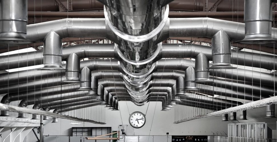 Индустриален интериор с метални тръби и стенен часовник, осветлен от естествена светлина.